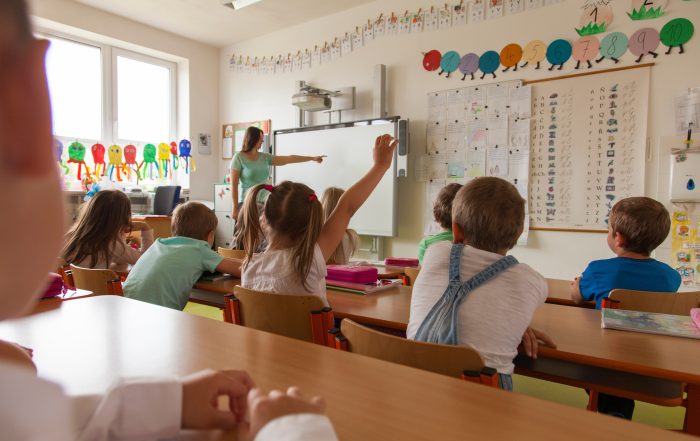 Lehrerin zeigt auf die interaktive Tafel. Grundschüler lernen und eine Schülerin meldet sich